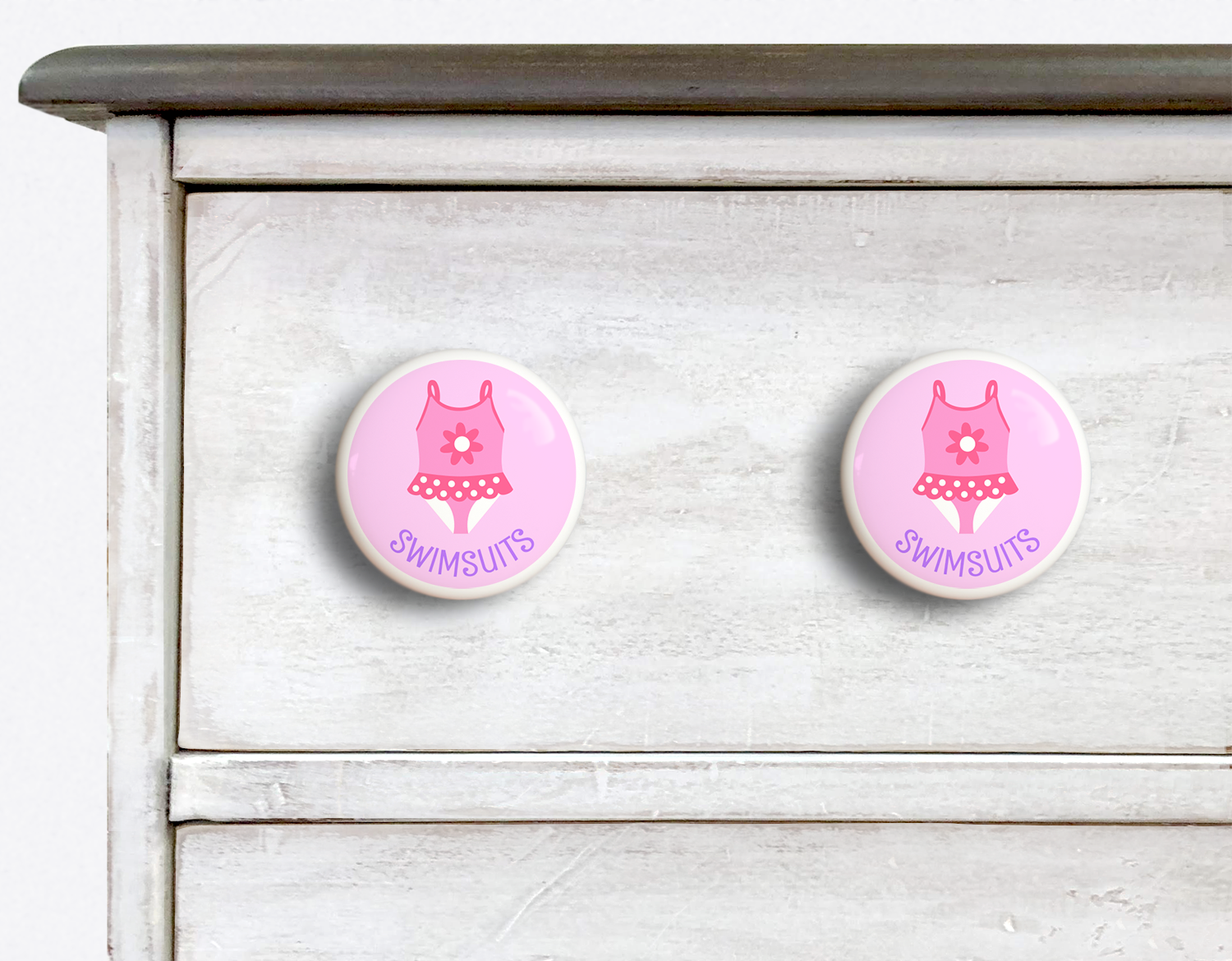 2 Ceramic drawer knobs on a dresser, Girl's bathing suit on a pink ground with the word Swimsuit written below