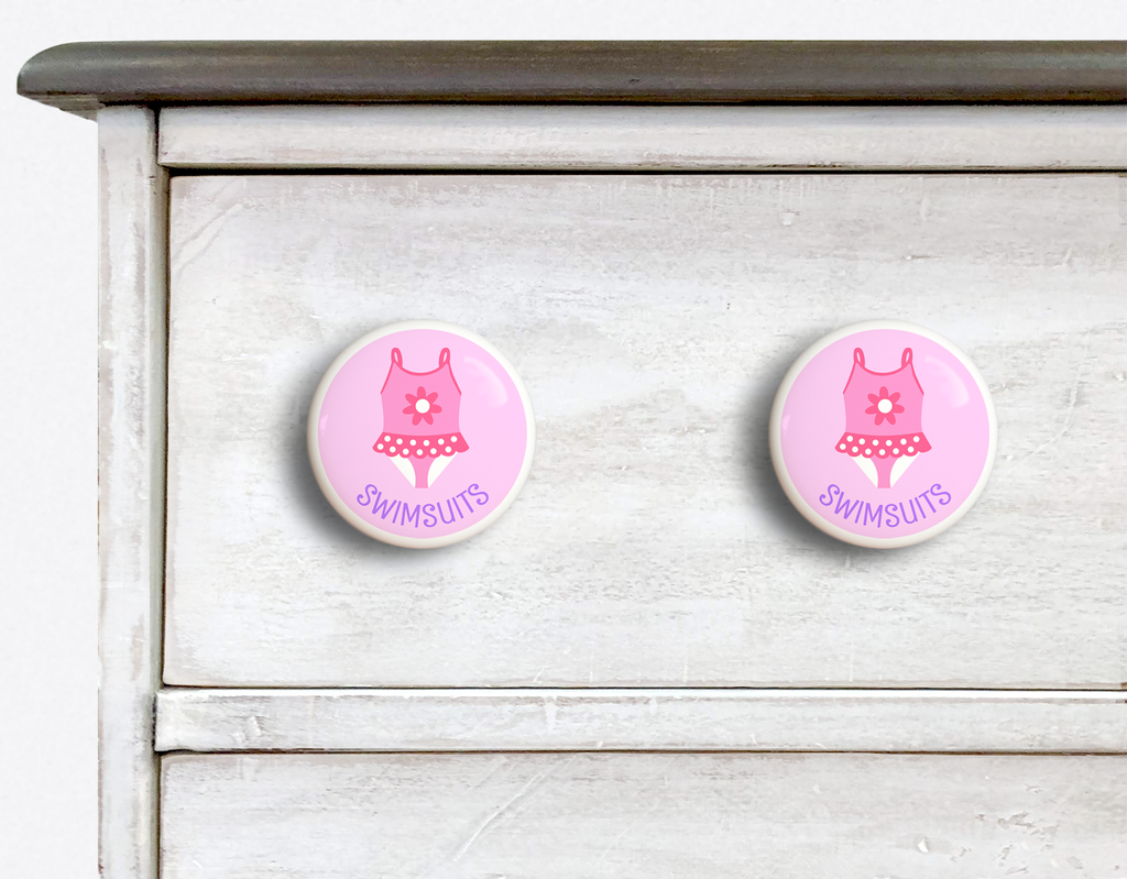 2 Ceramic drawer knobs on a dresser, Girl's bathing suit on a pink ground with the word Swimsuit written below