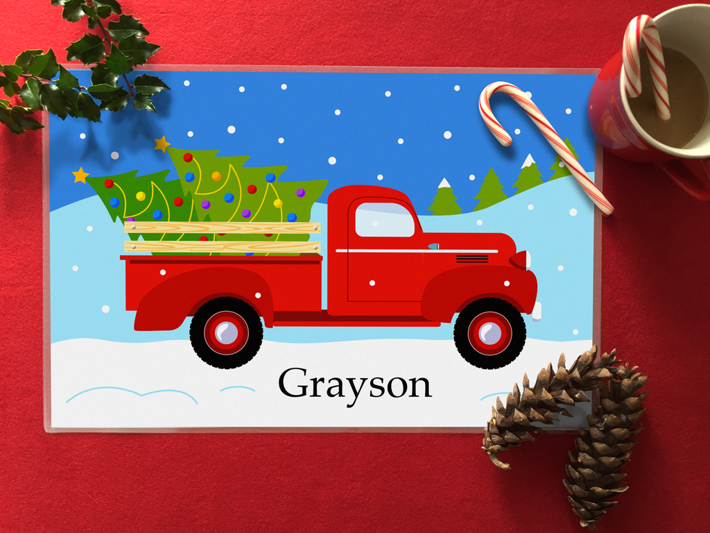 Christmas kids personalized placemat with vintage red pickup truck, christmas trees and snow with child's name at the botton. Placemat is on a red table top with pinecones, holly and hot chocolate.