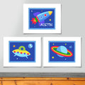 Set of 3 space themed prints, one of brightly colored rocketship and personalized with childs name, one of planet with rings and one of UFO, all on dark blue backgrounds with stars.