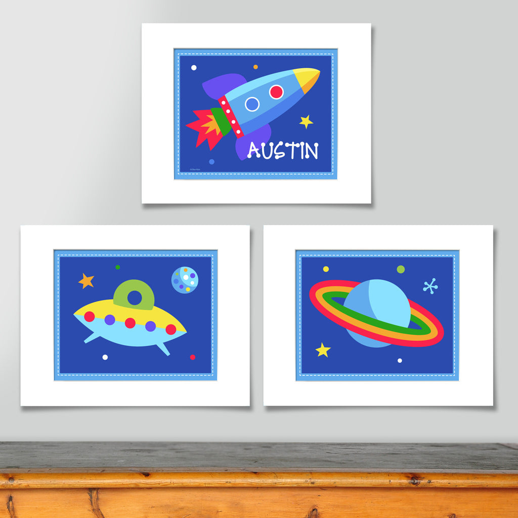 Set of 3 space themed prints, one of brightly colored rocketship and personalized with childs name, one of planet with rings and one of UFO, all on dark blue backgrounds with stars.